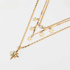 A gold layered necklace set featuring multiple star and bar charms, including one larger star pendant with a clear rhinestone and a small pearl accent, displayed on a clean white background.