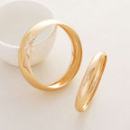 Two polished gold rings displayed on a light beige surface, with one standing upright and the other lying flat, next to a white ceramic bowl.