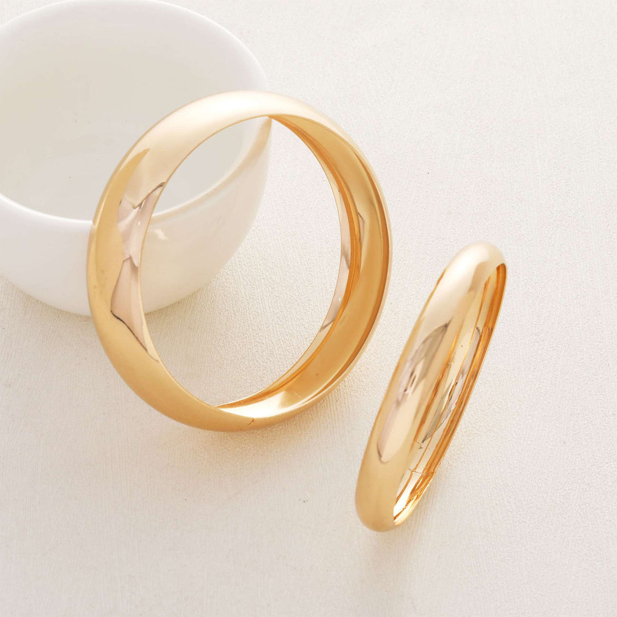 Two polished gold rings displayed on a light beige surface, with one standing upright and the other lying flat, next to a white ceramic bowl.