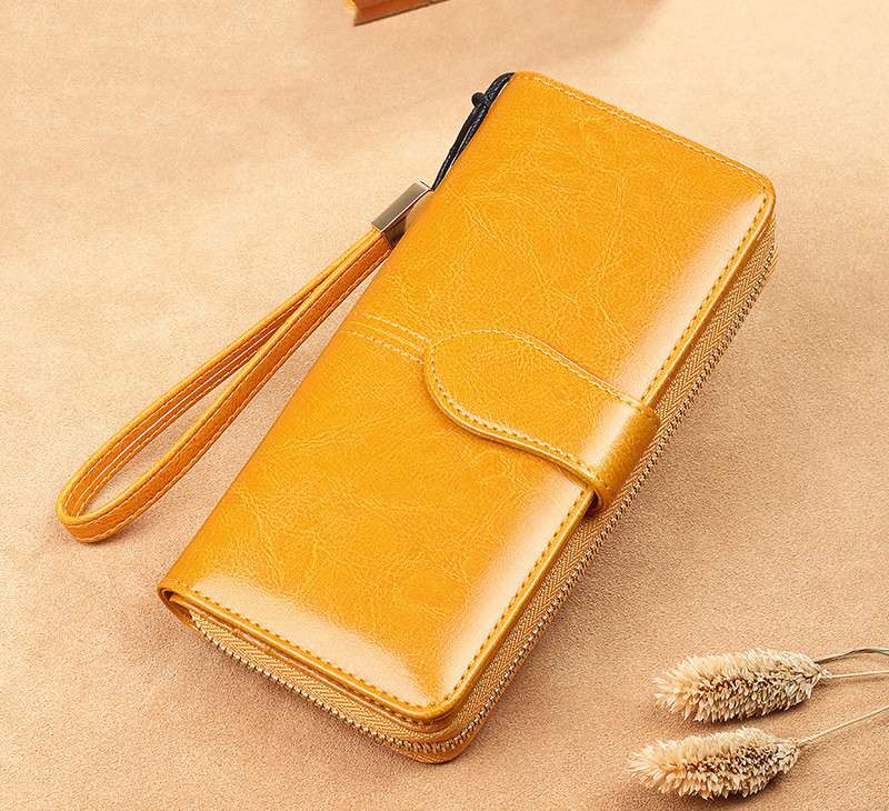 A mustard yellow faux-leather zip-around wallet with a wrist strap, shown on a textured beige background with dried decorative grass stems.