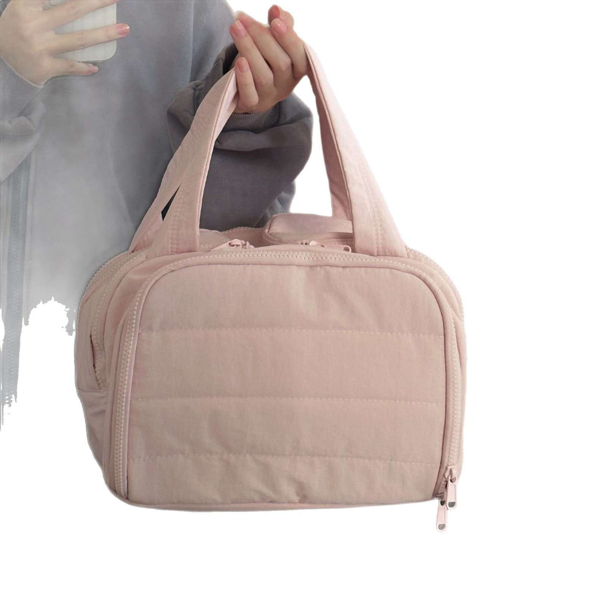 A light pink quilted nylon travel bag with dual top handles, shown being carried by a person whose hands and torso are partially visible on the left against a white background.