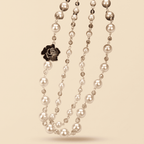 A multi-strand pearl necklace with gold-toned metal links, small faceted clear beads, and a black rose-shaped charm, displayed on a solid beige background.