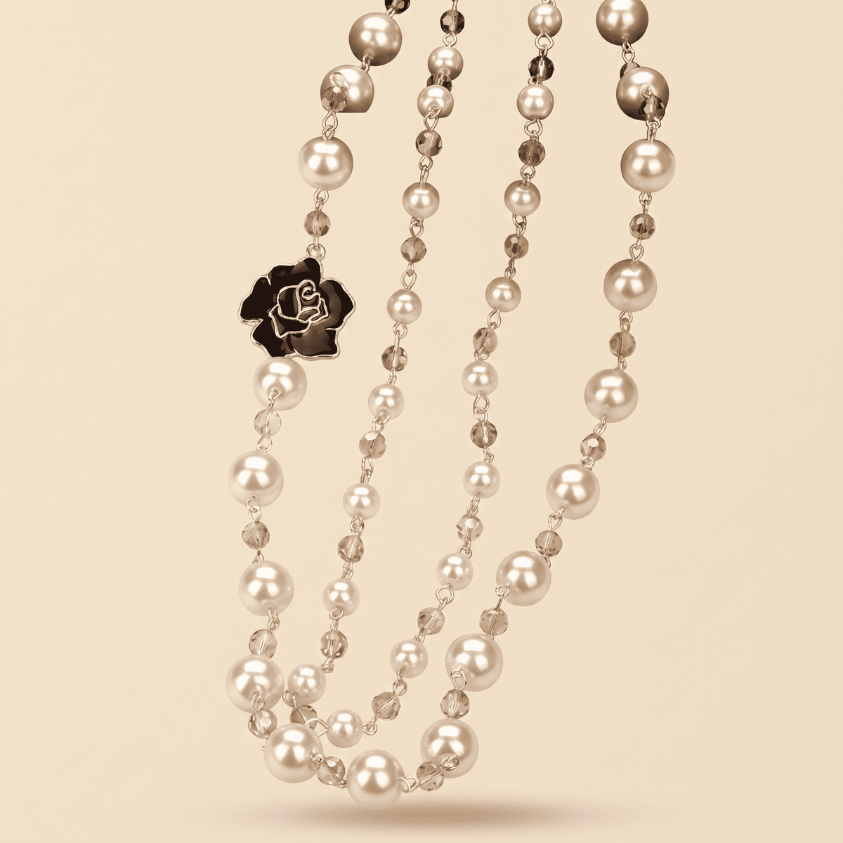 A multi-strand pearl necklace with gold-toned metal links, small faceted clear beads, and a black rose-shaped charm, displayed on a solid beige background.
