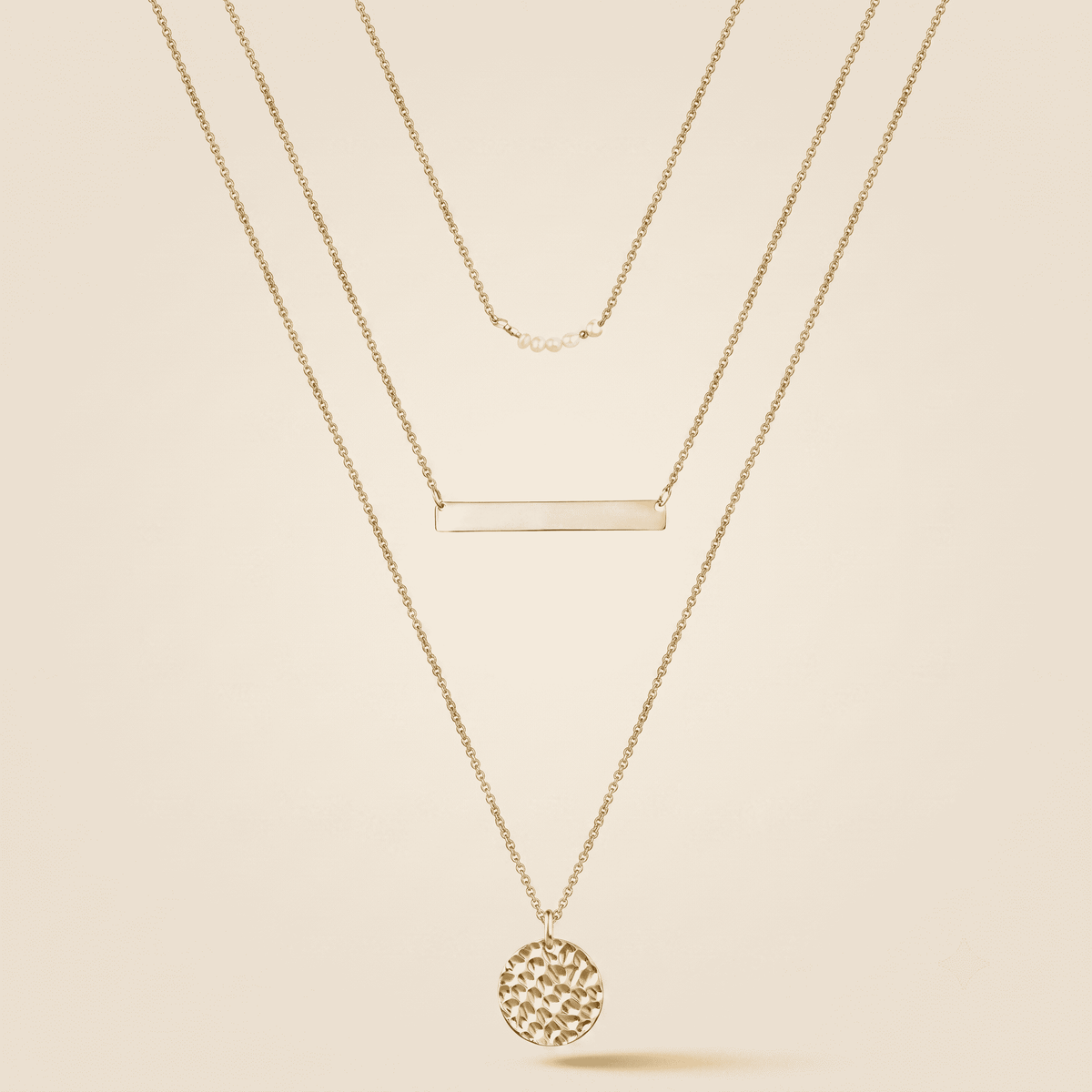 A gold layered necklace set featuring a delicate chain, a horizontal bar, and a textured circular pendant, displayed on a warm beige background.