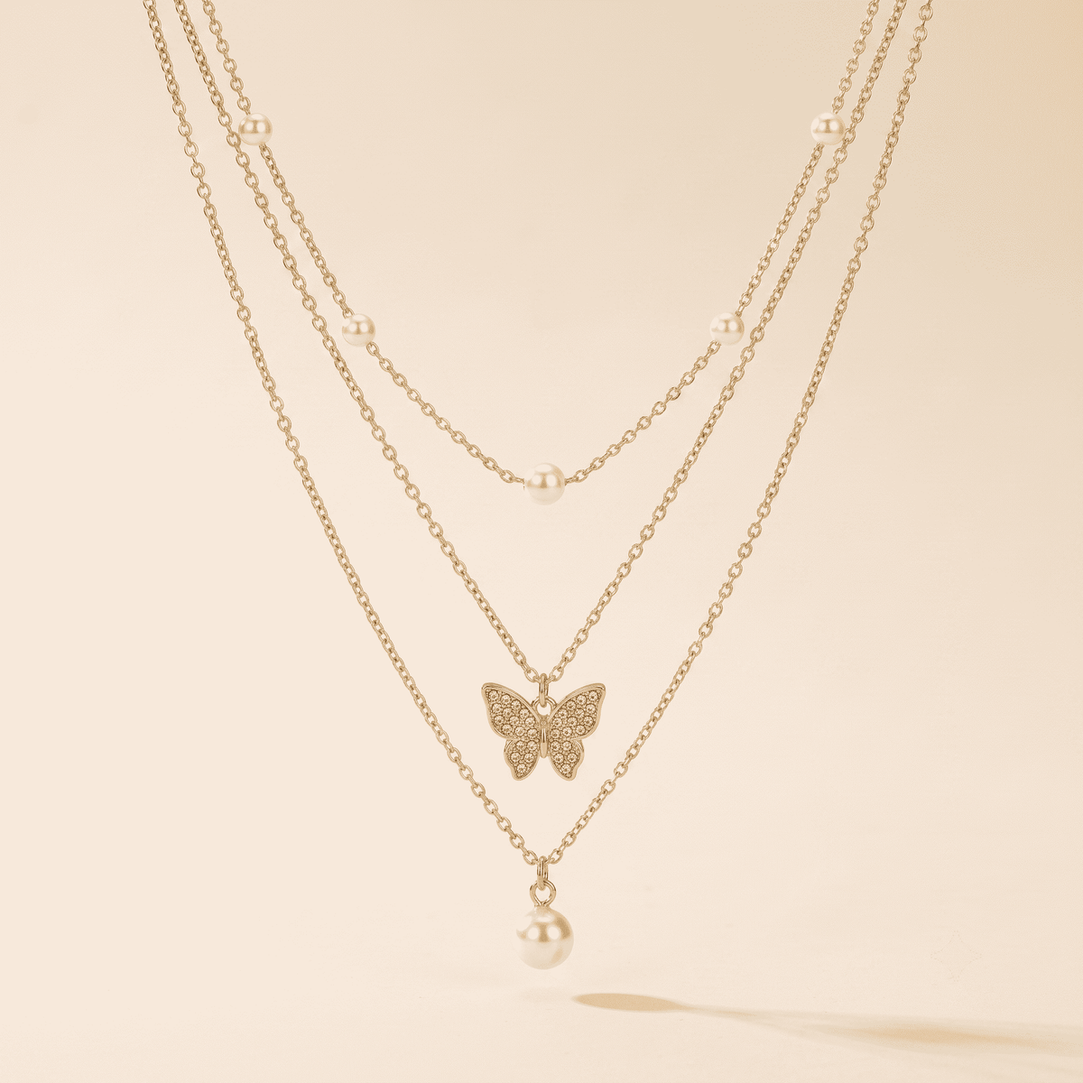 A delicate three-layer gold necklace featuring a central butterfly pendant paved with small clear stones and a dangling pearl bead, displayed against a warm cream background.