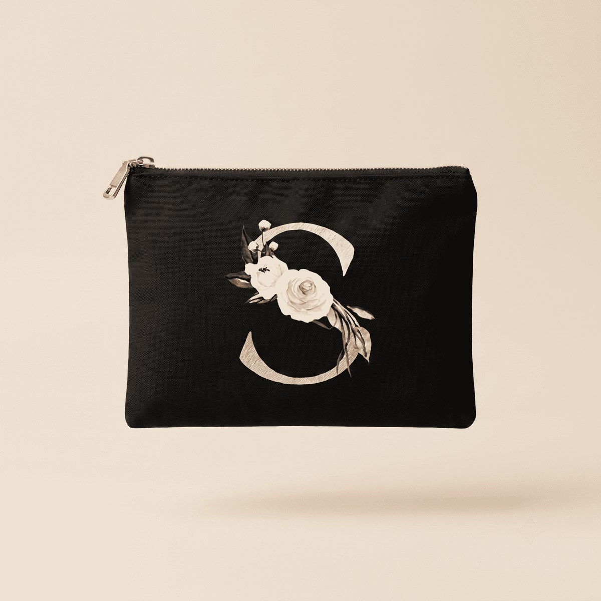 A black zippered pouch featuring a large decorative monogram letter 'S' embellished with white and beige flowers, set against a solid light beige background.