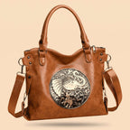 A brown leather handbag with a large circular silver-toned elephant emblem on the front, dual top handles, and an adjustable shoulder strap, shown on a plain beige background.
