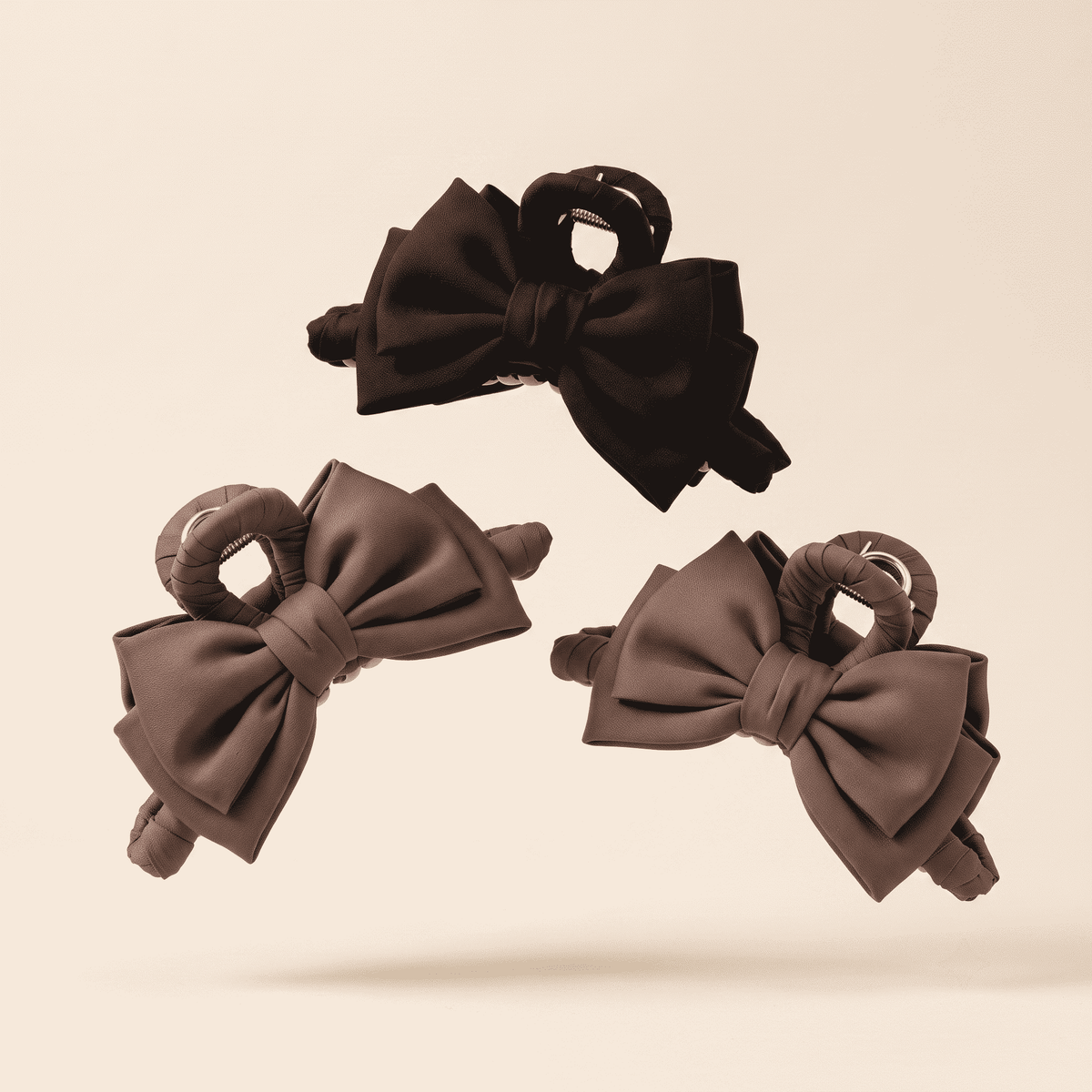 Three hair claw clips with large bow details, shown in black and taupe-brown tones against a solid light beige background.
