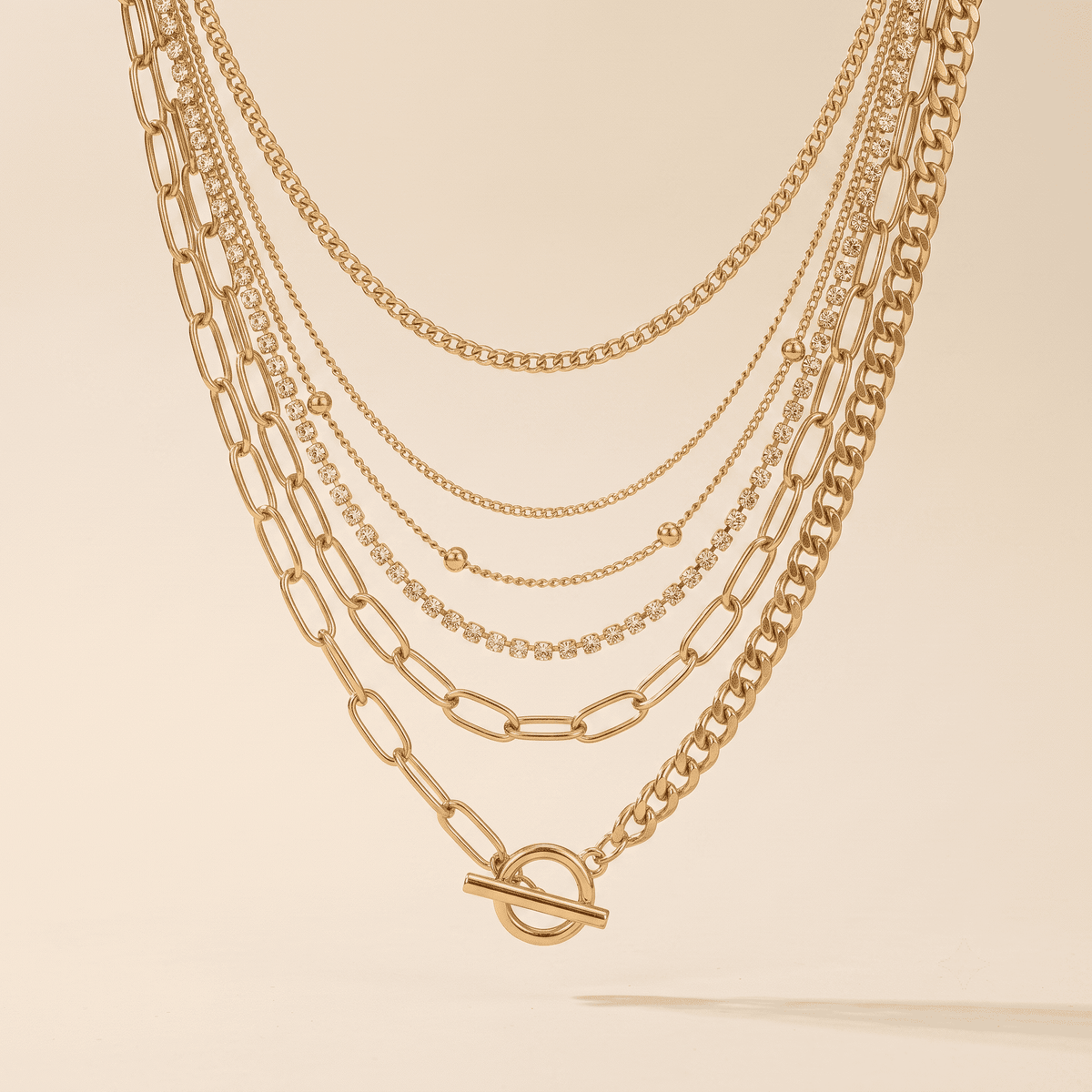 A layered gold necklace set featuring multiple chain styles including paperclip, figure-eight, cable, and delicate bead chains, with a toggle clasp on the longest chain, displayed against a warm cream background.