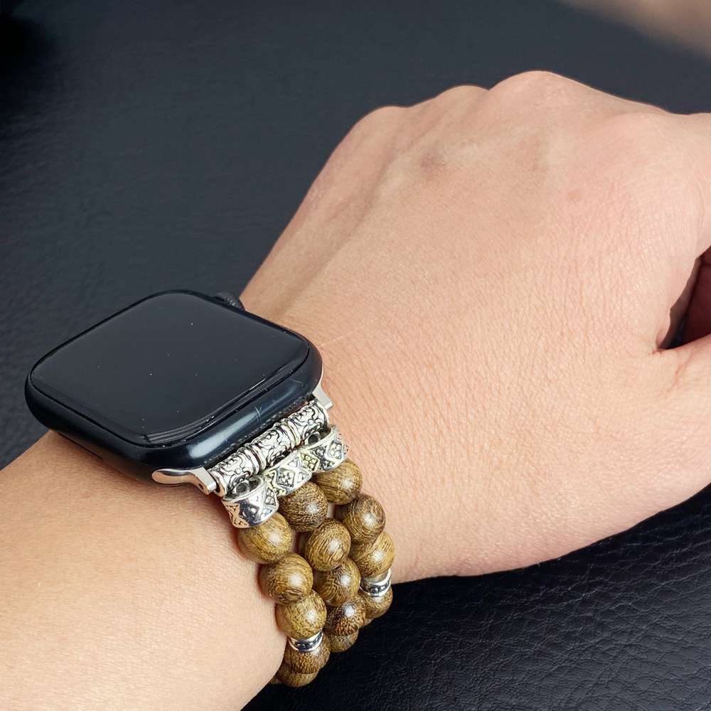 A close-up of a person's wrist wearing a black smartwatch with a decorative multi-strand bracelet featuring brown wooden beads and ornate silver-toned metal accents.