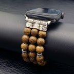A close-up of a smartwatch with a decorative band made of round brown wooden beads and ornate silver-toned metal links, resting on a black textured surface.
