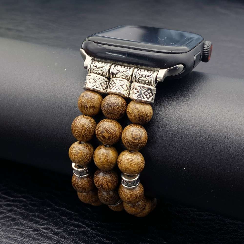 A close-up of a smartwatch with a decorative band made of round brown wooden beads and ornate silver-toned metal links, resting on a black textured surface.