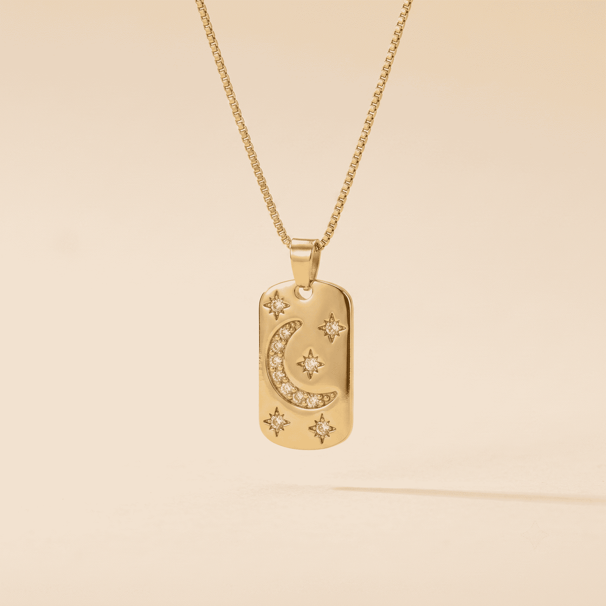 A gold dog tag necklace featuring a crescent moon and star design, embellished with small clear stones, shown on a matching gold chain against a warm beige background.