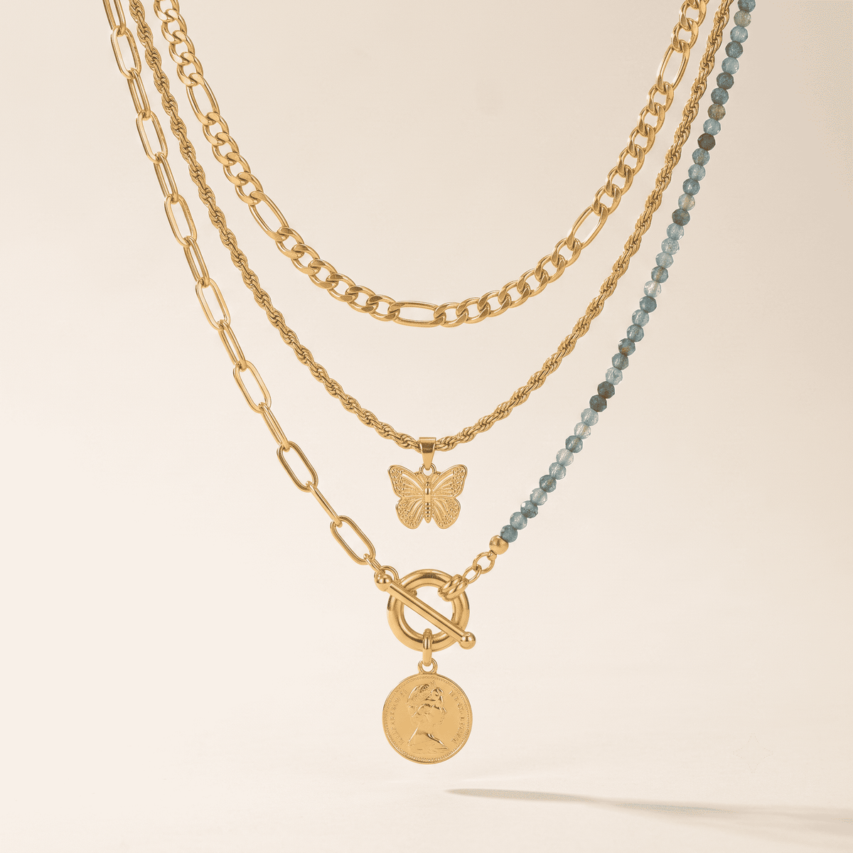 A layered gold necklace set featuring a butterfly pendant, a round coin charm, and a beaded strand of light blue beads, displayed on a warm cream background.