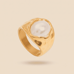 A gold ring featuring a large, smooth white pearl in a bezel setting with a textured, organic gold outline, shown against a solid beige background.