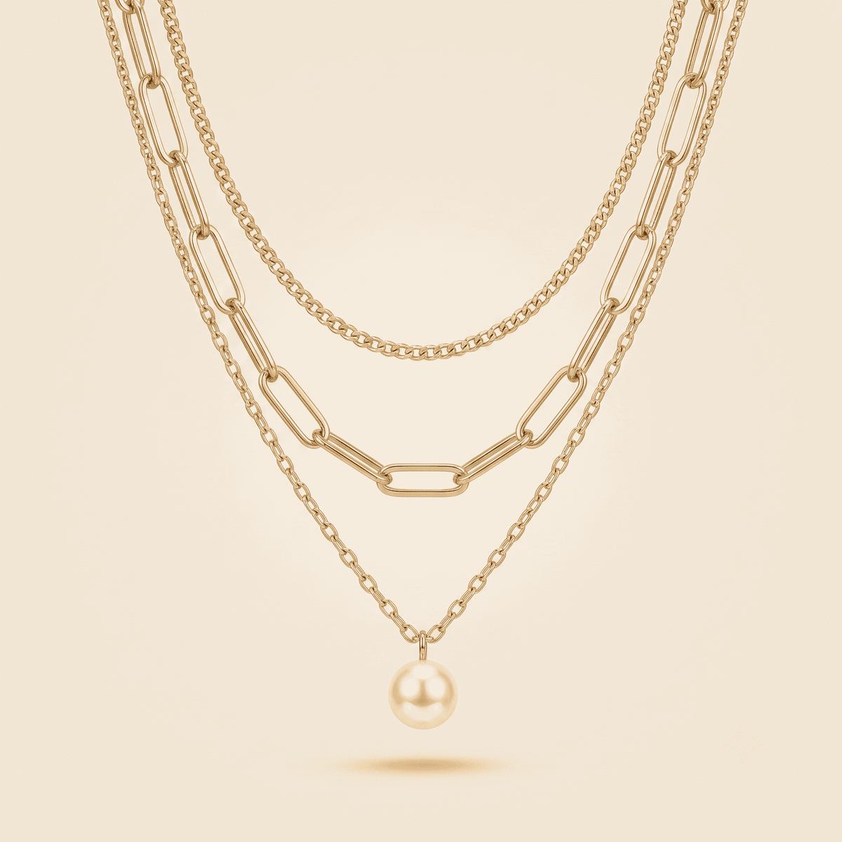 A layered gold necklace set featuring three cascading chain styles and a single dangling pearl pendant, displayed on a warm beige background.