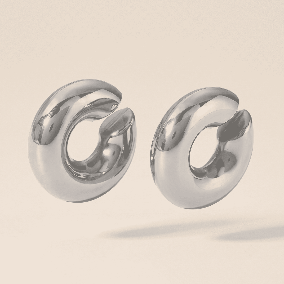 A pair of chunky, polished silver hoop earrings with a unique open, curved design, shown on a warm beige background.