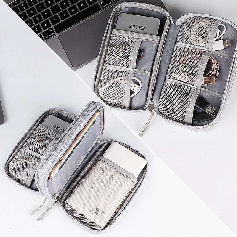 Gray zippered organizer case shown open in two views on a white surface, with a laptop corner visible in the top left. The case has mesh pockets, elastic loops, and compartments holding cables, cards, and small accessories.