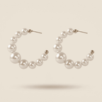 A pair of open hoop earrings made of white faux pearls of varying sizes, connected by small gold-toned beads, on a light beige background.