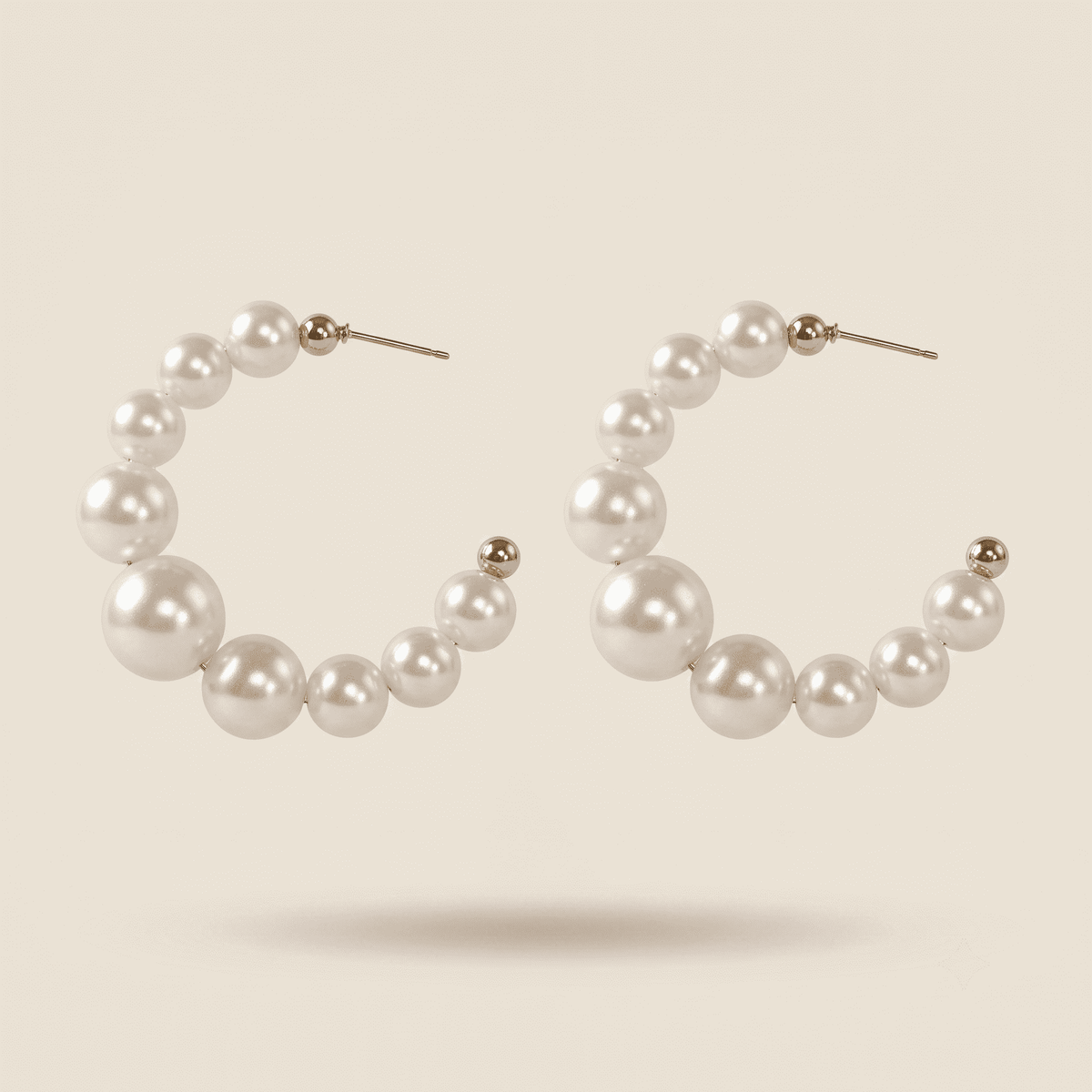 A pair of open hoop earrings made of white faux pearls of varying sizes, connected by small gold-toned beads, on a light beige background.
