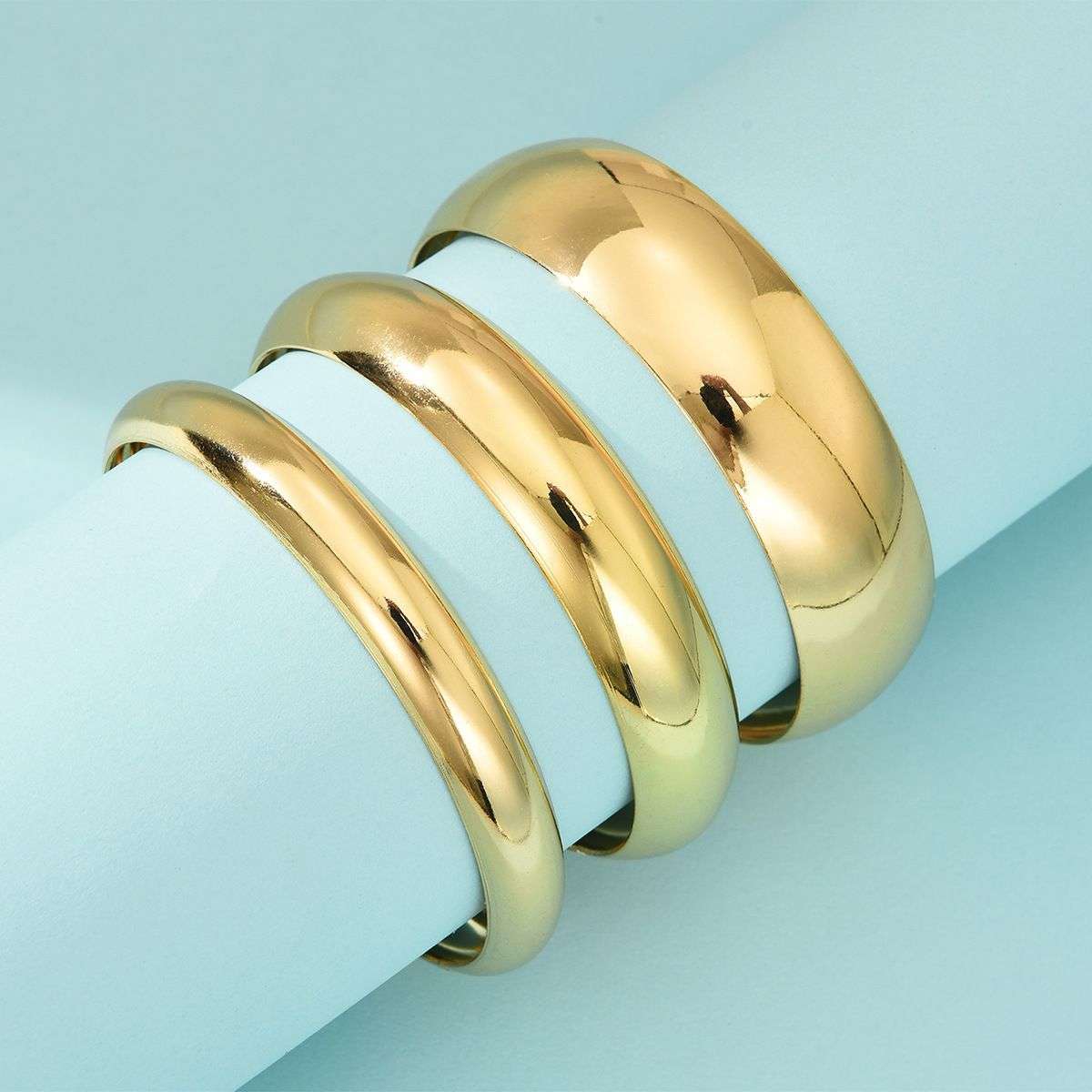 Three polished gold metal bands, likely wedding or fashion rings, stacked diagonally on a light blue cylindrical display prop.