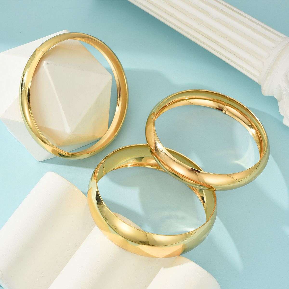 Three polished gold-toned hoop earrings displayed on white geometric blocks against a light blue background.