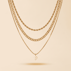 Three layered gold-tone necklaces displayed against a warm beige background. The top two layers feature chunky chain styles, while the bottom layer is a delicate chain with a single small pearl pendant.