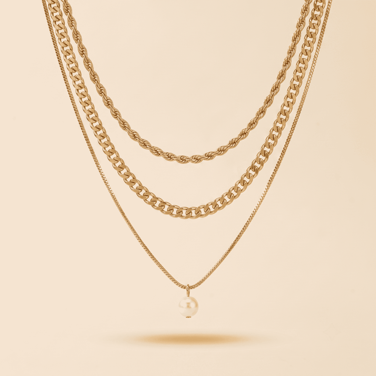 Three layered gold-tone necklaces displayed against a warm beige background. The top two layers feature chunky chain styles, while the bottom layer is a delicate chain with a single small pearl pendant.